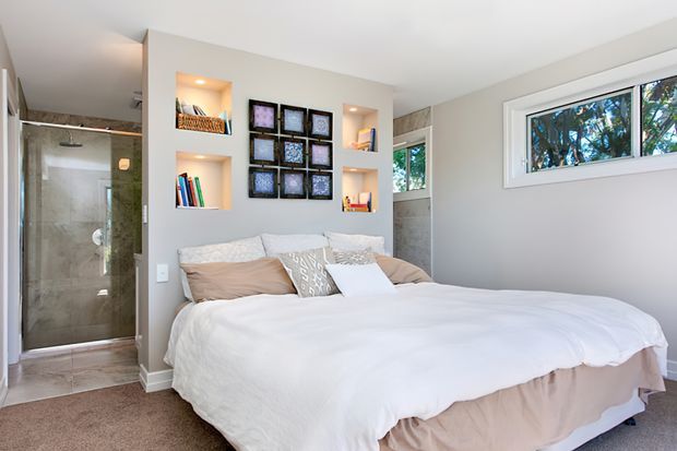 Bright, compact bedroom with a large neatly made bed, built-in illuminated display niches, framed wall art, and a tiled glass-enclosed shower visible through an opening at the left. Neutral finishes, soft bedding, and natural light create a clean, comfortable atmosphere.