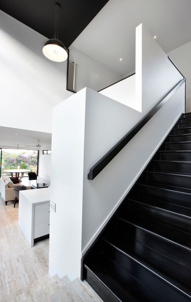 Bright contemporary residential stairway with glossy black timber stairs and matching dark handrail, set against crisp white walls and a dramatic black ceiling plane. The stair landing overlooks an open-plan dining and living area with a white kitchen island, while recessed lights and a spherical pendant provide illumination. Pale timber flooring, minimalist joinery, and large glazing create a high-contrast, modern interior.
