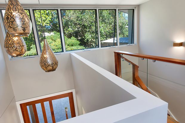 A bright upper-level stair landing with white walls, a white half-height stairwell balustrade, timber handrails and posts, and a large run of black-framed windows overlooking leafy greenery. Decorative patterned pendant lights hang over the stair void, while a timber-and-glass stair railing and a timber-framed door are visible below.