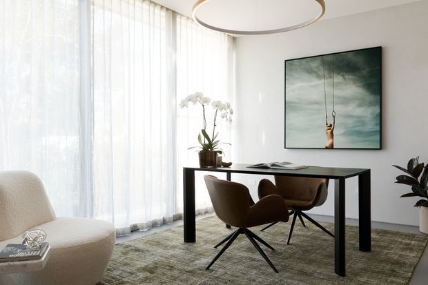 A bright, refined home office with floor-to-ceiling sheer curtains, a dark rectangular desk, two brown upholstered swivel chairs, a textured neutral rug, and a sculptural circular pendant light. A large framed artwork and indoor plants add visual interest, while a curved lounge chair occupies the foreground.