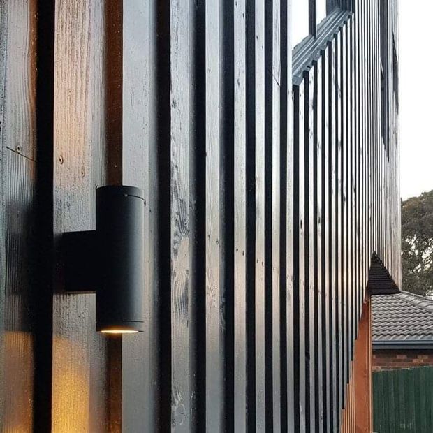 Close-up exterior architectural detail showing a dark vertically ribbed metal facade with alternating projecting and recessed strips, a black cylindrical wall light, and a contrasting timber-toned vertical panel. Part of a tiled-roof brick building and greenery are visible in the background.