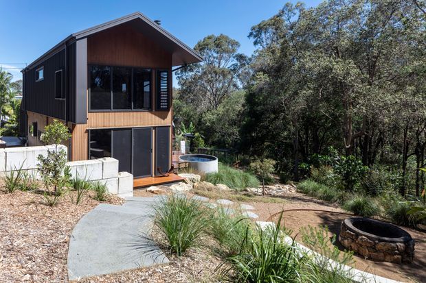 Exterior view of a compact two-storey dark-clad house set within a natural bushland landscape. The building has a steep gabled roof, warm timber detailing beneath the gable, large dark-framed windows and doors, and a concrete pathway leading through landscaped native planting. A small circular stone-edged fire pit or garden feature is visible in the foreground.