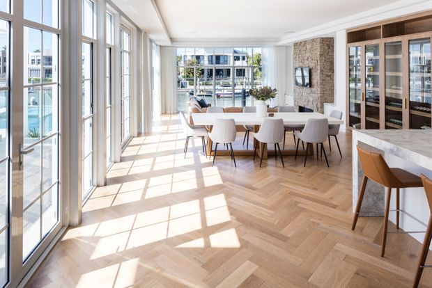 Bright, refined open-plan dining and entertaining area with extensive floor-to-ceiling glazing along one side, a large dining table with upholstered chairs, pale herringbone timber flooring, a natural stone feature wall, and built-in glass-front cabinetry. A marble or light stone kitchen island with timber stools is visible in the foreground. The room has abundant daylight, views toward a waterfront or pool-like exterior, and a sophisticated contemporary coastal feel.