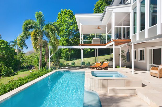 Bright exterior view of a contemporary multi-level residence with extensive white-framed glazing, open balconies and covered outdoor living areas overlooking a landscaped sloping site. A swimming pool and raised spa dominate the foreground, with palm trees and lush planting around the property.