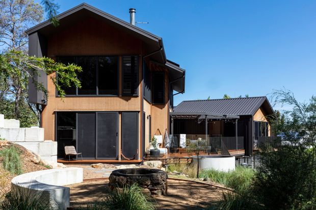 Exterior view of a contemporary multi-level timber-clad home on a landscaped sloping site. The house has a prominent pitched gable roof, dark window and door frames, extensive glazing, a covered outdoor entertaining area, and a landscaped courtyard with a circular fire pit.