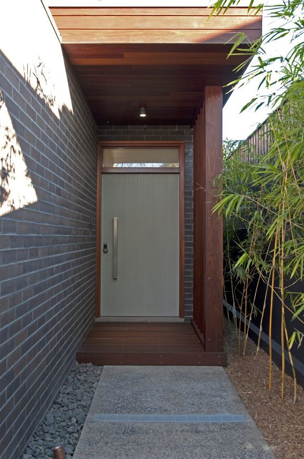 A narrow, landscaped residential entrance walkway leading to a contemporary front door. The entry is framed by dark charcoal brickwork, warm timber decking and a timber-lined projecting canopy supported by a vertical timber post. The ribbed pale-grey door has a long vertical pull handle and a narrow glazed transom above. Bamboo planting and a dark screening fence line the right side, while exposed dark stone provides drainage edging along the brick wall.
