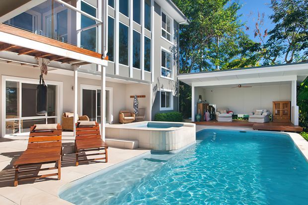 A bright contemporary multi-storey residence arranged around a large swimming pool. The image shows extensive floor-to-ceiling glazing, a covered patio with timber sun loungers, a raised square spa, and an open poolside garage or cabana containing seating and storage. White and pale grey exterior surfaces contrast with warm timber decking and joinery, with mature trees surrounding the property.