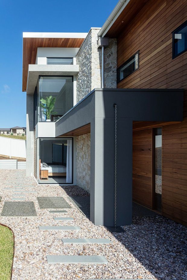 A contemporary multi-level residential facade viewed from an exterior walkway. The building combines warm horizontal timber cladding and soffits, rough natural stone walls, smooth dark grey rendered structural elements, white rendered volumes, and large dark-framed windows. A gravel landscape with rectangular concrete stepping stones leads toward a recessed timber entry door. A partially visible bedroom can be seen through ground-floor glazing.
