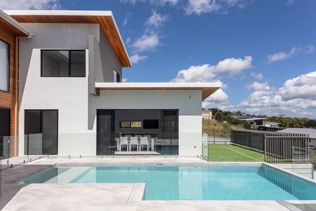 Contemporary multi-level residence with white rendered walls, warm timber soffits and accents, dark window frames, and a flat-roofed projecting volume. The rear outdoor area includes a large turquoise swimming pool, pale paved terrace, glazed balustrade, and fenced lawn, with neighboring homes visible beyond.