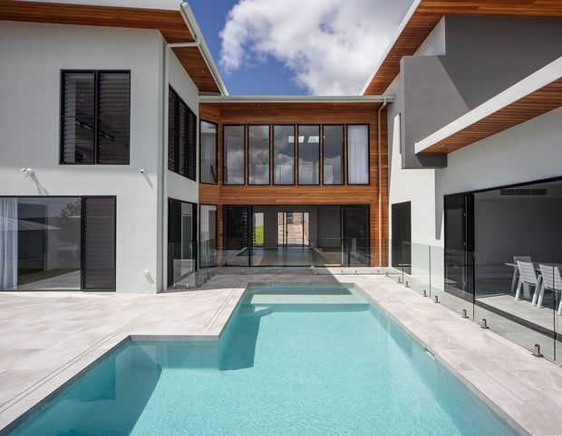 Contemporary multi-wing residence arranged around a private swimming pool courtyard. The home features white rendered walls, dark-framed glazing and louvres, warm timber soffits, a central glazed upper-level connection, and a sheltered outdoor dining area beside the pool.