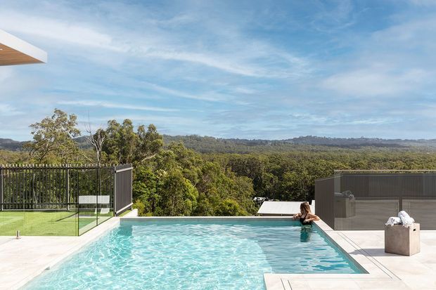 A bright elevated outdoor pool terrace overlooking a dense forested landscape. The rectangular turquoise pool is bordered by pale paving, dark metal privacy screens, and a modern flat-roofed structure, with a person seated at the pool edge. The scene has a contemporary architectural character and expansive treetop views.