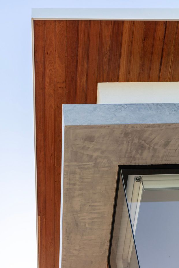 Close-up architectural detail showing a warm reddish-brown vertical timber-clad wall or soffit beside a pale grey textured concrete or cement-finished surface and dark-framed glazing. The image is tightly cropped and appears to focus on exterior facade materials and junction detailing.