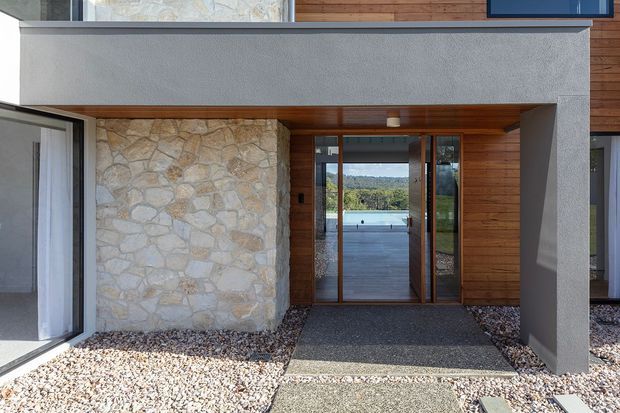 Contemporary residential entrance with a sheltered recessed doorway, natural irregular stone feature wall, warm horizontal timber cladding and soffit, and smooth grey rendered concrete forms. Glazed double doors open toward a deck and pool or landscape beyond, with gravel landscaping and concrete stepping surfaces at the threshold.