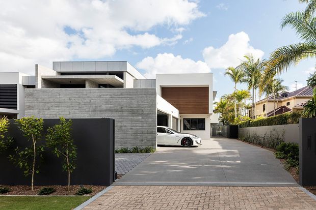 Street-level exterior view of a contemporary multi-level residence with a broad concrete driveway and paved frontage. The home features strong geometric volumes, exposed grey concrete walls, crisp white rendered elements, dark metal screening and gates, and a prominent projecting upper volume with a warm timber-lined soffit. A covered carport accommodates a vehicle, while palms and hedges frame the property.