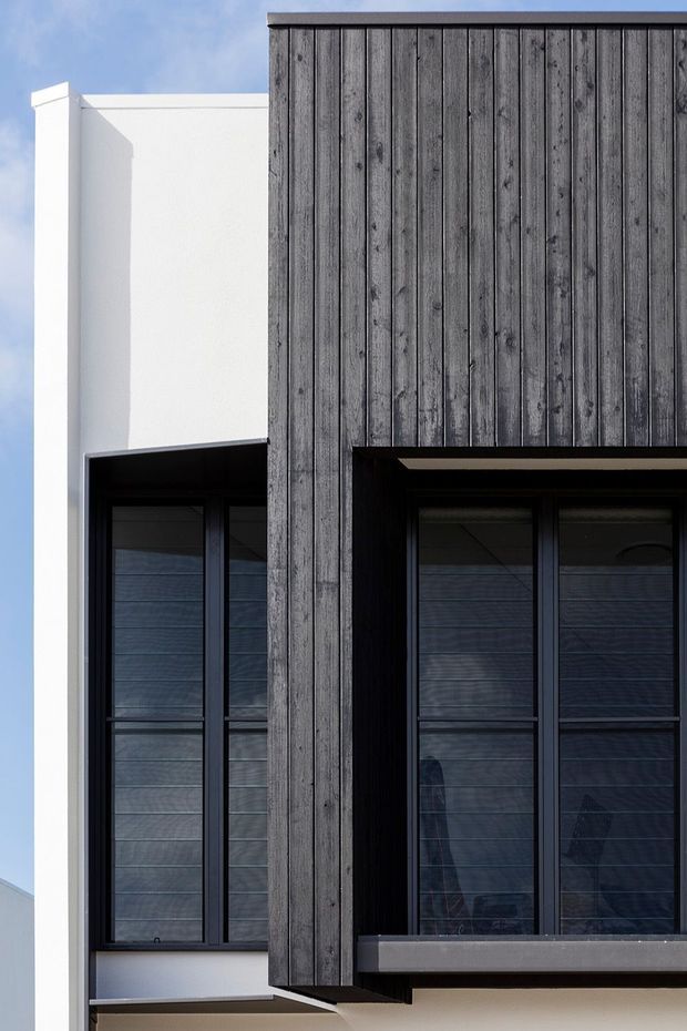 Close-up exterior view of a contemporary building facade, combining dark vertically boarded timber cladding with crisp white rendered wall sections and deeply recessed black-framed windows. The composition highlights strong geometric forms, layered projections, and contrasting light and dark materials.