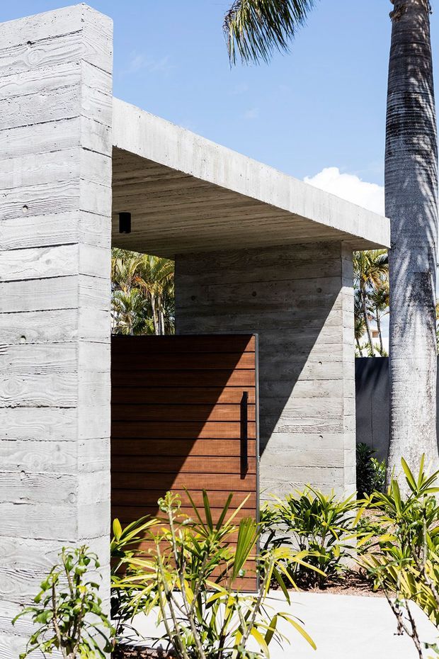 Exterior entry gate framed by a raw, board-formed concrete structure with a broad flat canopy. A dark timber slatted gate, minimalist black hardware, tropical planting, and a palm tree create a contemporary tropical entrance.