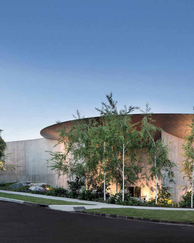 Exterior architectural view of a sculptural, curved contemporary building set behind landscaped planting. The structure features a broad copper-toned sweeping roof or canopy, pale vertical wall cladding, warm concealed lighting, and a gently curving pedestrian path beside a dark paved street.