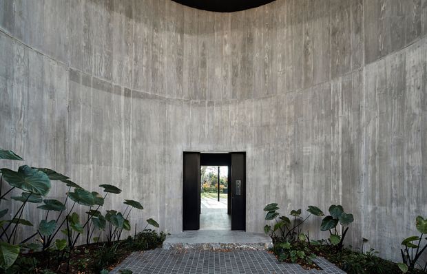 A curved, monolithic concrete entryway with tall vertically textured concrete walls, a black-framed doorway, and a small tiled forecourt bordered by lush tropical planting. The view through the doorway reveals an exterior path beyond.