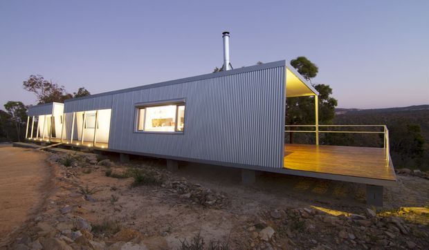 A compact elevated contemporary cabin or dwelling set on a rocky, sparsely vegetated site at dusk. The long rectangular structure has dark grey corrugated metal cladding, a projecting timber deck with simple metal balustrades, large glazed openings, and a bright yellow recessed outdoor area beneath a sloping roof element. A flue projects from the roof, and the building appears supported above the ground on piers.