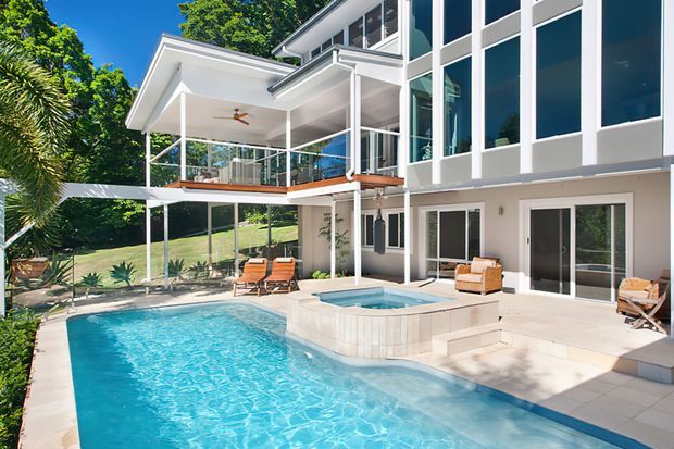 Bright exterior view of a multi-level contemporary residence with extensive floor-to-ceiling glazing, white rendered walls, covered balconies and terraces, and a large swimming pool with an elevated square spa in the foreground. The house is set against lush greenery on a gently sloping site, with outdoor lounge furniture and tropical planting around the pool.