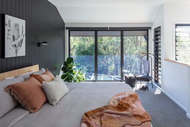 Bright contemporary bedroom with a large upholstered bed, layered neutral and rust-toned bedding, a dark vertical-panel feature wall, and extensive floor-to-ceiling glazing overlooking leafy surroundings. The room includes a potted plant, lounge chair, wall artwork, and carpeted floor, with strong natural light and clean modern detailing.