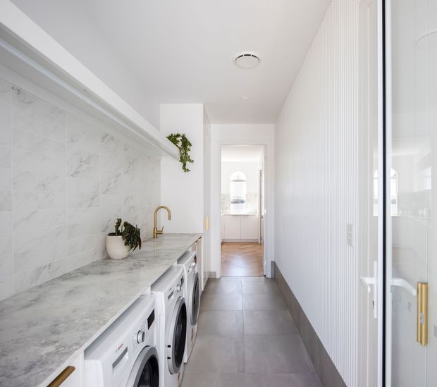 Bright, narrow contemporary laundry room with a long marble-look benchtop over front-loading appliances, white marble-look tiled splashback, white cabinetry and wall panelling, grey floor tiles, and a brass gooseneck tap. A glazed door and hallway lead toward a bright adjoining room.