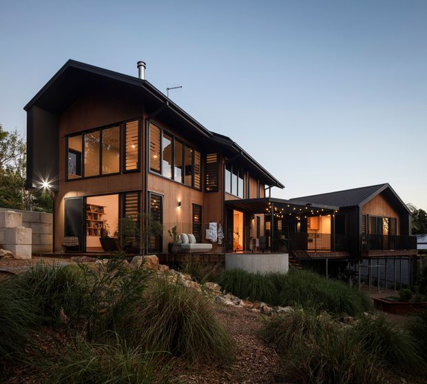 Exterior architectural view of a contemporary multi-level home on a landscaped, sloping site. The dwelling features dark timber or charcoal cladding, extensive black-framed glazing, pitched roofs, warm interior lighting, and a covered outdoor entertaining area with festoon lights. Native grasses, rocks, retaining walls, and steps form the foreground landscape.