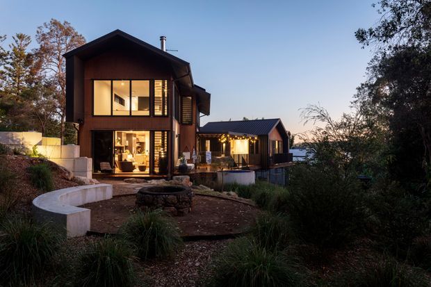 Dusk exterior view of a contemporary multi-level residence set among landscaped vegetation. The home features dark vertical timber cladding, large illuminated windows, projecting gabled roof forms, and an outdoor entertaining area with a curved built-in concrete seating wall and fire pit. Water is faintly visible beyond the house.
