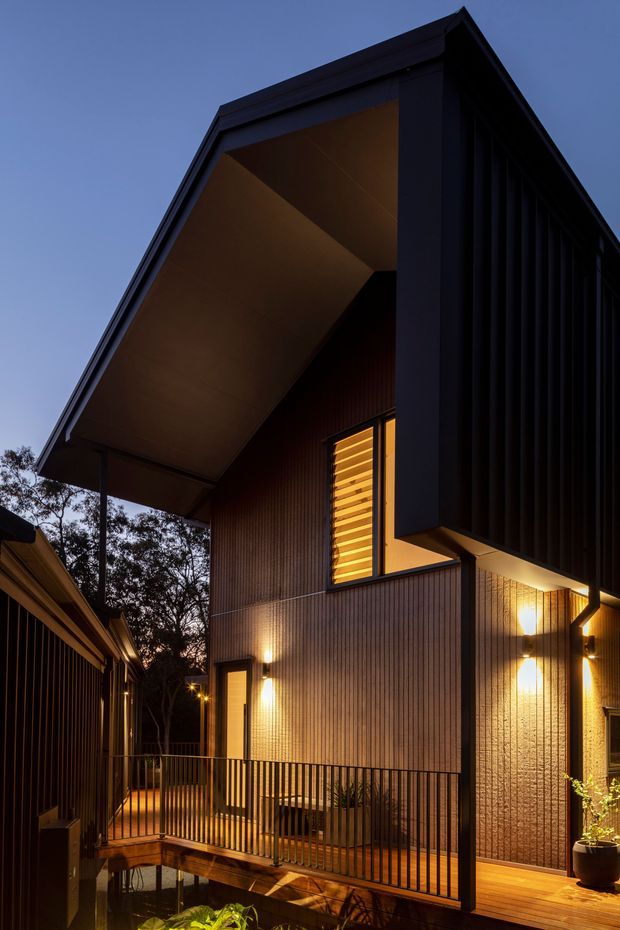 Dramatic dusk exterior view of a contemporary multi-level residence with a steep gabled roof, deep projecting eaves, dark vertical cladding, warm timber-toned wall surfaces, and illuminated windows and wall lights. A raised timber deck and narrow bridge with black metal balustrades connect the home to an adjacent structure, with trees and a small planted water feature below.