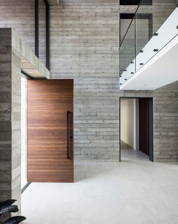 A minimalist double-height residential entryway with exposed horizontal-board concrete walls, a large horizontal timber pivot door, polished light concrete flooring, and a glass-balustraded upper-level walkway. A dark-framed interior doorway opens to a corridor, emphasizing the sculptural, open circulation space.