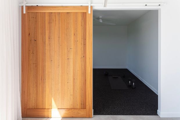 Interior view of a compact garage or utility space with a large vertical timber sliding barn door on white metal hardware. The open doorway reveals a simple room with white walls, dark carpet flooring, and a ceiling fan; the surrounding interior has white trim and a pale hard floor.