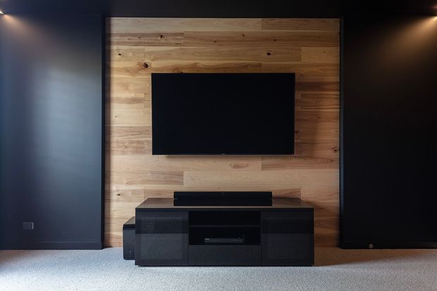 A dark, minimalist media wall featuring a wall-mounted flat-screen television set against a warm horizontal timber panel, with a low black media console below and tall dark acoustic or storage panels on either side. Recessed lighting creates a moody, high-contrast atmosphere, while pale carpet covers the floor.