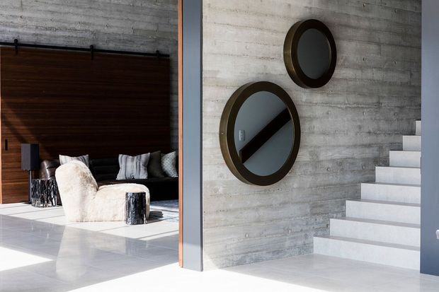 Contemporary interior showing a polished light floor, exposed textured concrete walls, a white open-riser staircase, and a partially visible bedroom or lounge area beyond a large dark timber sliding door. Two round dark-framed mirrors are mounted on the concrete wall, reflecting the room and stair area. The space has strong natural light, clean lines, and a restrained palette of white, grey, black, and dark wood.