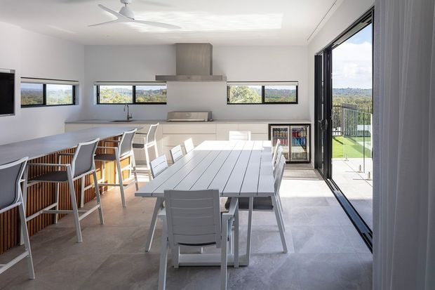 Bright contemporary open-plan kitchen and dining area with a long island bench, white cabinetry, integrated stainless-steel rangehood, bar stools, and a large dining table. Wide windows and fully opening glazed doors provide abundant daylight and views to the landscaped exterior.
