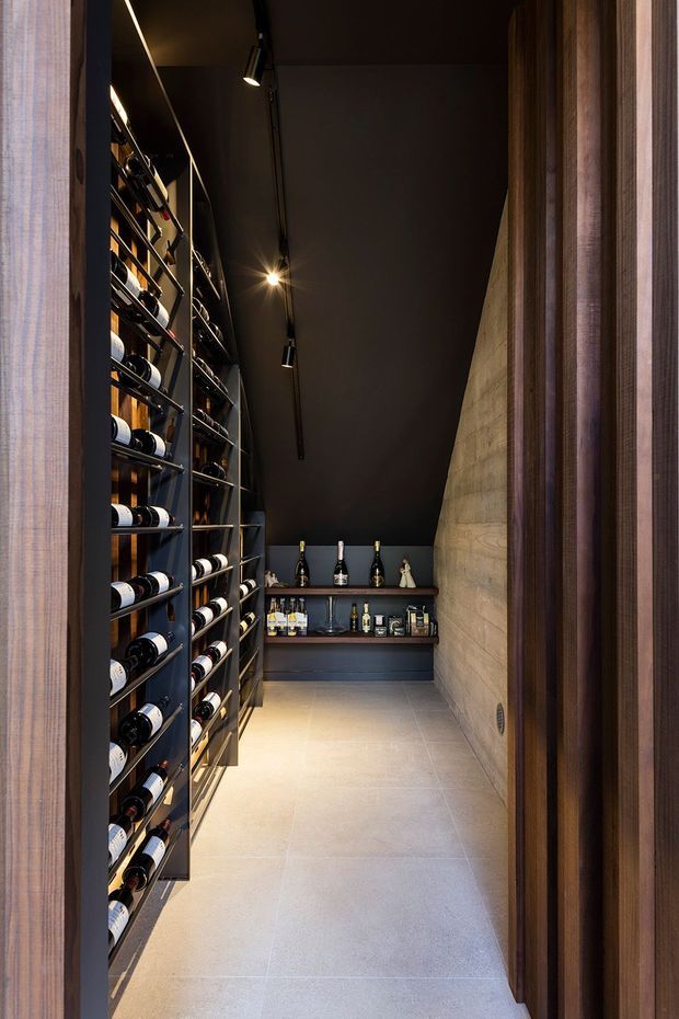 A narrow, purpose-built wine storage room viewed through a timber-framed opening. Floor-to-ceiling dark metal wine racks line the left wall, while a dark built-in bar and bottle display occupy the far end. The right wall has a raw, textured concrete finish, with a dark ceiling and track lighting creating a dramatic, warm atmosphere. Large pale floor tiles contrast with the dark joinery and walls.