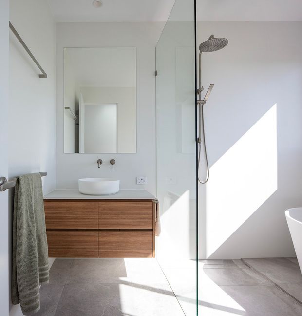 Bright contemporary bathroom with a floating timber vanity, white vessel basin, wall-mounted taps, frameless glass walk-in shower, and a partially visible freestanding bathtub. Large-format light grey floor tiles and white walls create a clean, minimalist setting, with strong sunlight casting geometric shadows across the room.