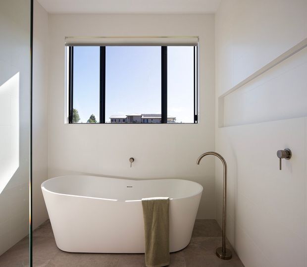 Bright contemporary bathroom featuring a freestanding white soaking bathtub, brushed bronze floor-mounted filler, matching wall-mounted controls, and a large black-framed horizontal window. The space has white walls with subtle decorative panel moulding and a warm grey tiled floor.
