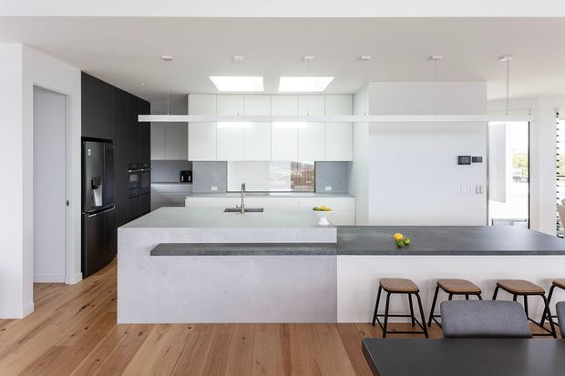 Bright contemporary open-plan kitchen with a large pale stone island and integrated breakfast bar, extensive white cabinetry, dark feature cabinetry around the appliances, timber flooring, and minimal pendant lighting. A dining table and chairs are visible in the foreground, with daylight entering from the adjacent living area.