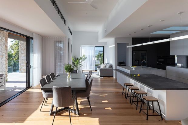 Bright open-plan contemporary kitchen, dining and living area with warm timber flooring, expansive glazing, a dark stone kitchen island, white cabinetry, and a long dining table. The room opens to an outdoor terrace through sliding glass doors and receives abundant natural light.