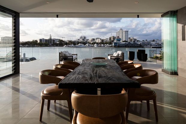 A refined waterfront dining area with a large sculptural dark timber dining table surrounded by curved brown leather chairs. Floor-to-ceiling glazing and open glass doors frame views across the water toward a dense coastal urban skyline. The room has pale stone or porcelain floor tiles, a smooth light ceiling, minimalist detailing, and strong indoor-outdoor connection.