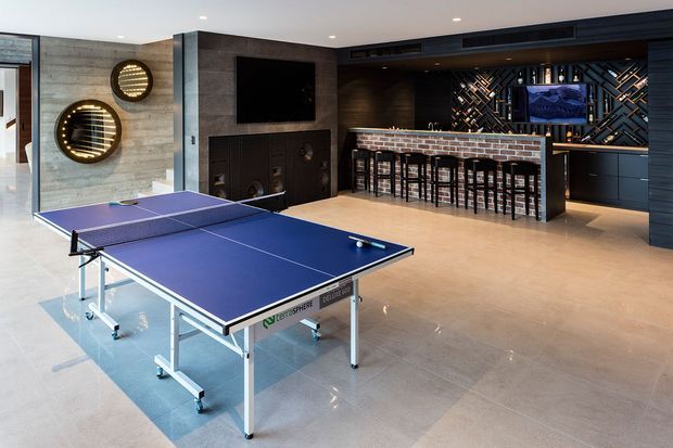 A spacious, high-end entertainment room featuring a full-size table tennis table in the foreground and a built-in home bar at the rear. The room has polished large-format flooring, dark wood and stone-look wall finishes, a wall-mounted television, integrated speakers, decorative circular wall lighting, and a patterned illuminated backdrop behind the bar.