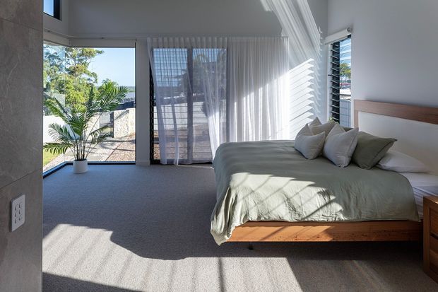Bright contemporary bedroom with a low timber platform bed, muted sage bedding, white walls, sheer full-height curtains, louvred windows, and expansive glazing overlooking greenery. The room has a soft carpeted floor and strong natural sunlight creating patterned shadows.
