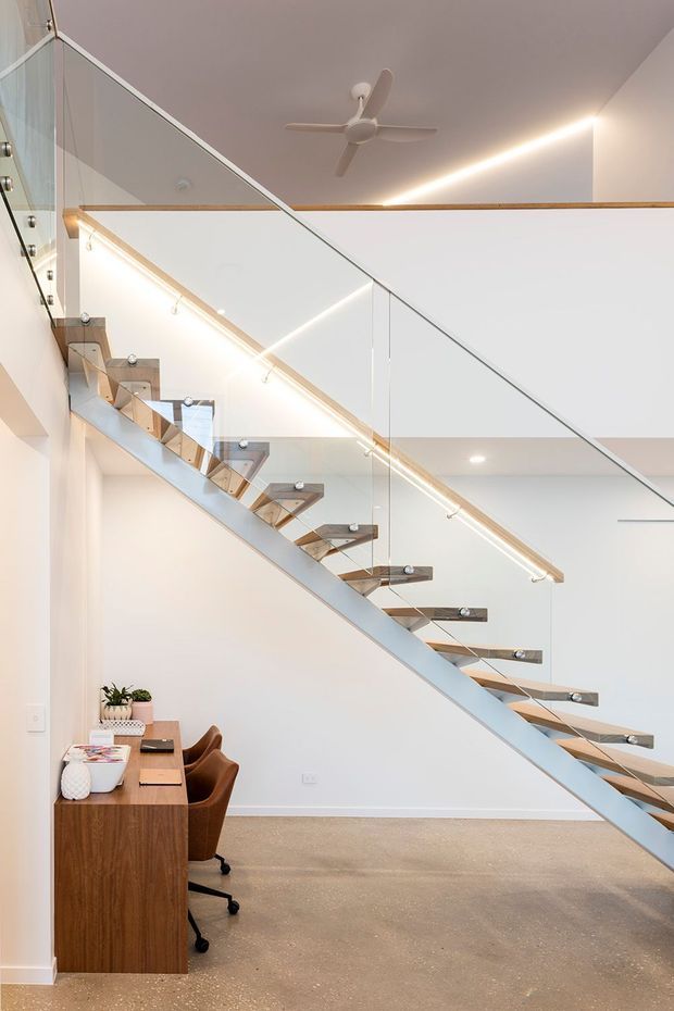 Bright contemporary interior featuring an open-riser floating staircase with timber treads, a pale structural stringer and clear glass balustrades. A compact timber desk with two office chairs sits beneath the stairs, while linear LED lighting, white walls and a ceiling fan create a clean, airy double-height circulation space.