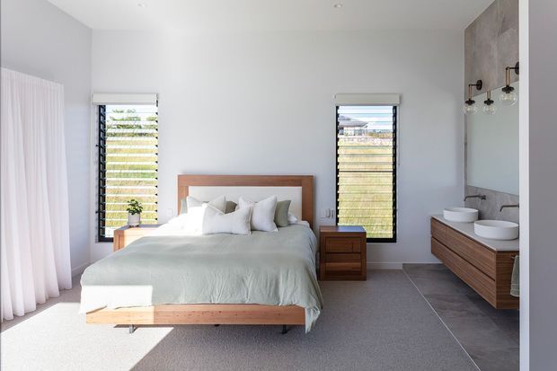 Bright contemporary bedroom with a timber bed frame and matching bedside tables, white bedding, pale walls, louvered windows, soft curtains, and an open timber vanity with twin vessel basins in an adjoining bathroom area. Grey tiled flooring defines the bathroom zone while the bedroom has a light carpet.
