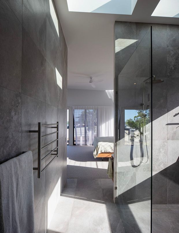 A contemporary bathroom with large-format dark grey stone-look wall and floor tiles, a frameless glass walk-in shower, wall-mounted shower fittings, and a heated towel rail. The bathroom opens directly onto a bedroom visible beyond, with a bed, carpeted floor, sheer curtains, and abundant natural light from skylights.
