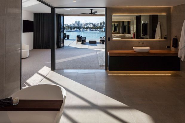 A contemporary open-plan bathroom and bedroom suite with a freestanding bathtub in the foreground, a floating dark timber vanity with a round vessel basin and illuminated mirror, and large sliding glass doors opening to a waterfront balcony. Neutral stone-look surfaces, dark cabinetry, recessed lighting, and strong sunlight create a refined resort-like atmosphere.