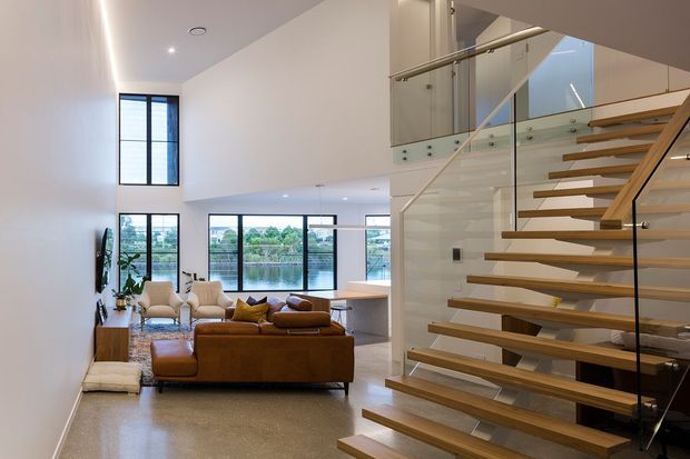 A bright contemporary open-plan living area viewed from a floating staircase. The room has a double-height volume, extensive black-framed glazing overlooking water, a warm brown sectional sofa, and a compact kitchen or bar area beyond. The staircase features open timber treads, a dark structural stringer, and clear glass balustrades with metal handrails. Smooth light walls and a polished neutral floor create an airy, refined interior.