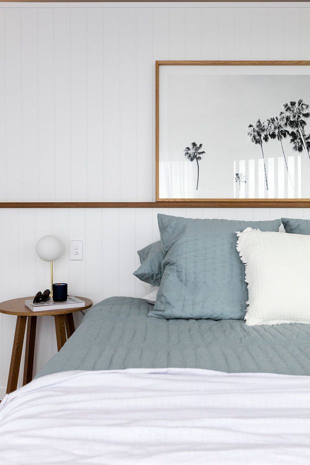 A bright, compact bedroom featuring a bed dressed in muted blue-grey textured bedding with a white fringed accent pillow. White vertical wall panelling is framed by warm timber trim, and a large timber-framed artwork or mirror displays silhouetted palm trees. A small round timber bedside table holds a spherical lamp and personal items.