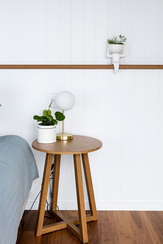 A bright, neatly styled bedroom corner featuring a small round timber side table beside the bed, a brass table lamp with a spherical white shade, and potted plants. The room has white vertically panelled walls with a slim timber picture ledge, warm timber flooring, and a soft blue-grey bed covering. The overall look is simple, fresh, and understated.