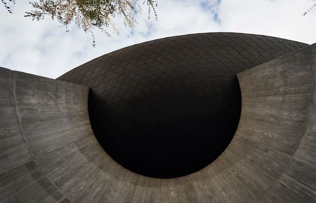 A close-up architectural exterior view of a dramatic curved concrete structure forming a deep, dark circular opening. Above the opening is a rounded, shingle-patterned dark roof or shell, while a small amount of sky and overhanging foliage is visible at the top edge. The composition is sculptural, minimal, and heavily focused on geometric form and material contrast.